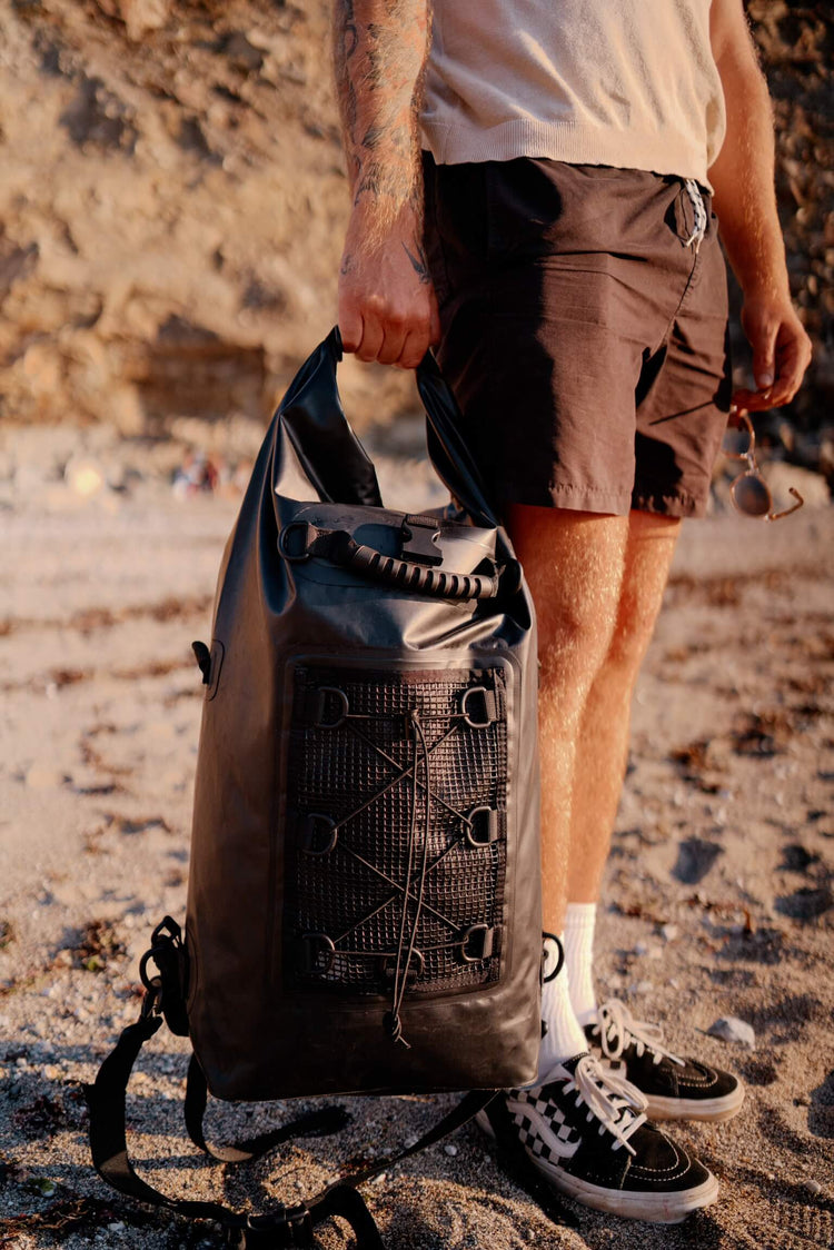 a man standing on the beach holding the black 30 litre salty kit dry back backpack 