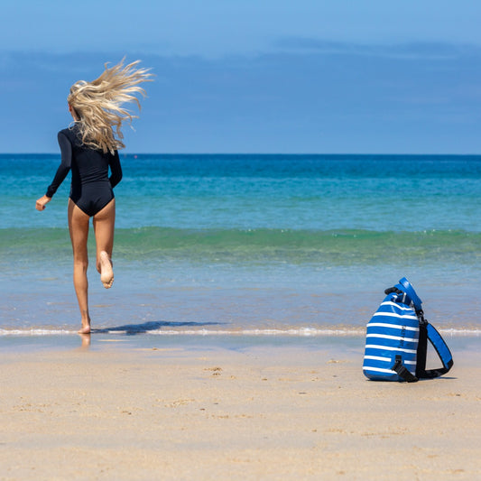 A woman in a black swimsuit runs barefoot on a sandy beach toward the ocean, with a blue and white striped backpack sitting on the sand nearby.