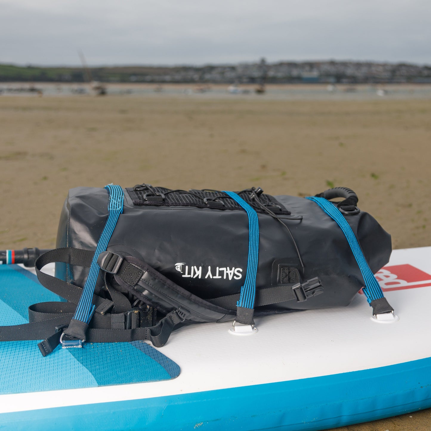 Black bag with blue straps on a blue and white paddleboard on a sandy beach.