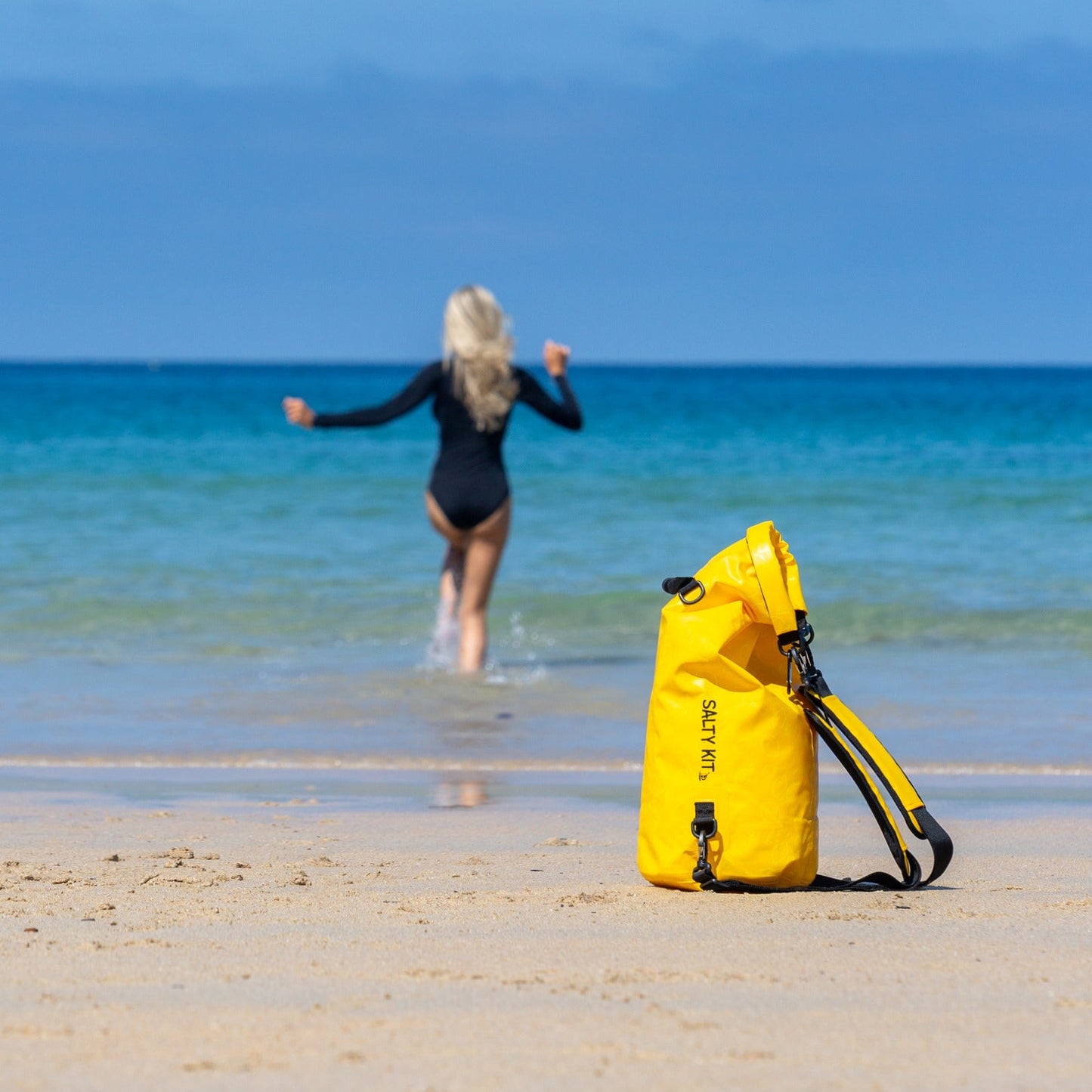 A bright yellow waterproof dry bag with lemon print and black straps sits on a sandy beach in the foreground, while a woman in a black swimsuit runs into the ocean water in the background under a clear blue sky.