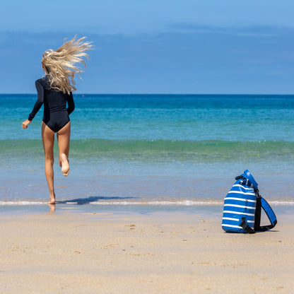 A woman in a black swimsuit runs barefoot on a sandy beach toward the ocean, with a blue and white striped backpack sitting on the sand nearby.