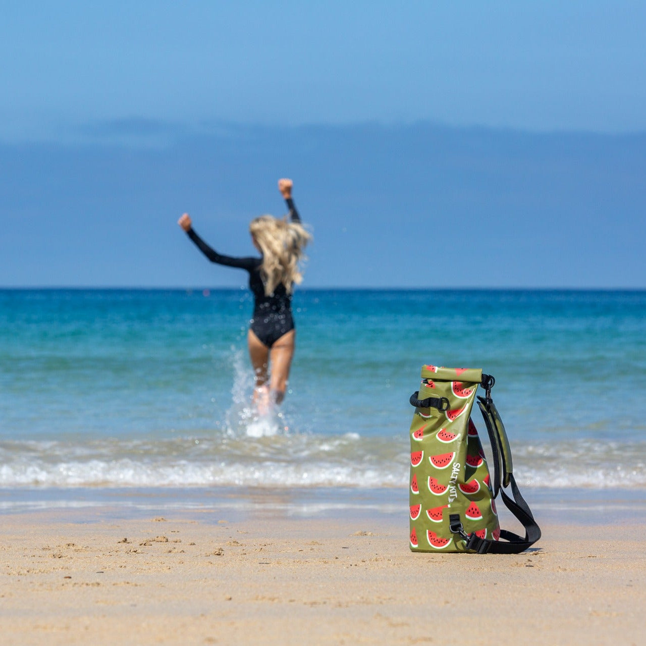 A watermelon dry bag with a watermelon print stands on a sandy beach in the foreground, while a woman in a black swimsuit runs into the ocean in the background under a clear blue sky.