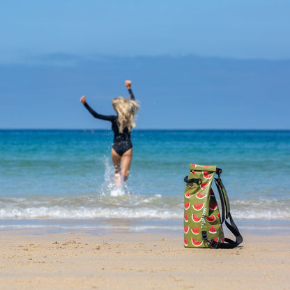 A watermelon dry bag with a watermelon print stands on a sandy beach in the foreground, while a woman in a black swimsuit runs into the ocean in the background under a clear blue sky.