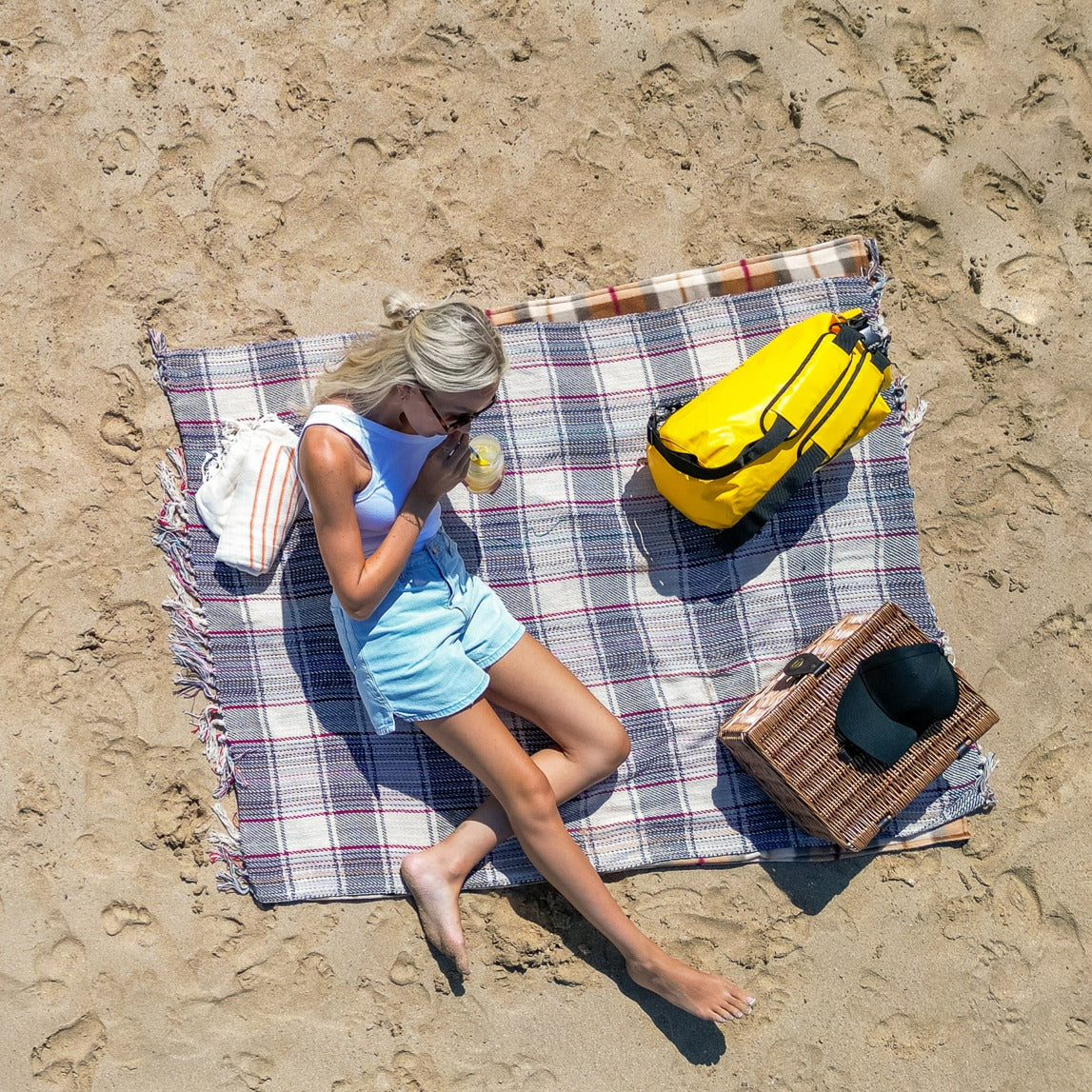 An aerial view of a woman sitting on a plaid beach blanket on sandy ground, drinking from a cup. A bright yellow waterproof bag, a striped towel, and a wicker basket with a black hat are placed around her.
