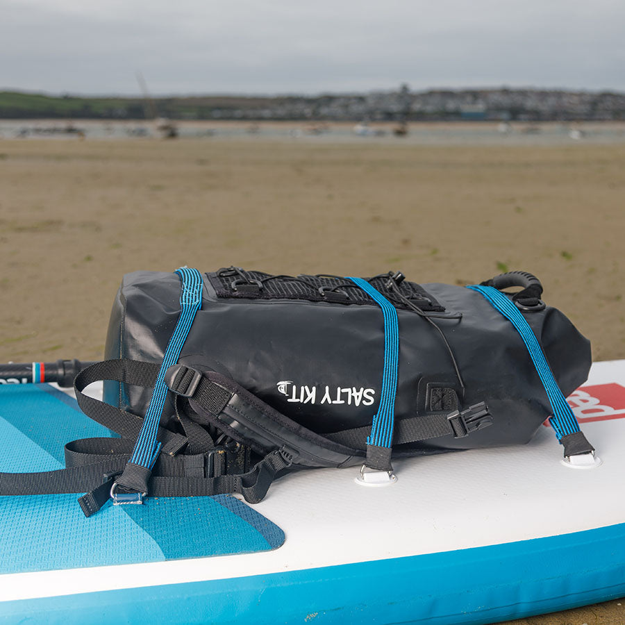 Black waterproof bag with blue straps on a blue and white paddleboard at a beach.