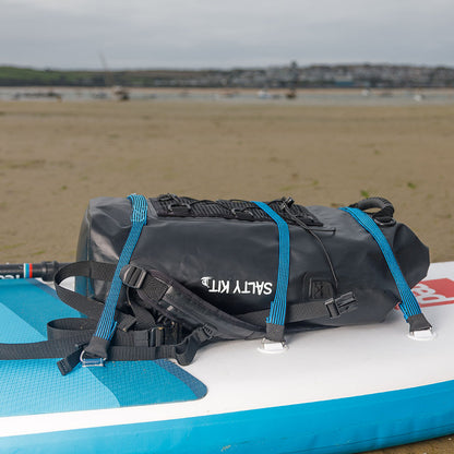 Black waterproof bag with blue straps on a blue and white paddleboard at a beach.