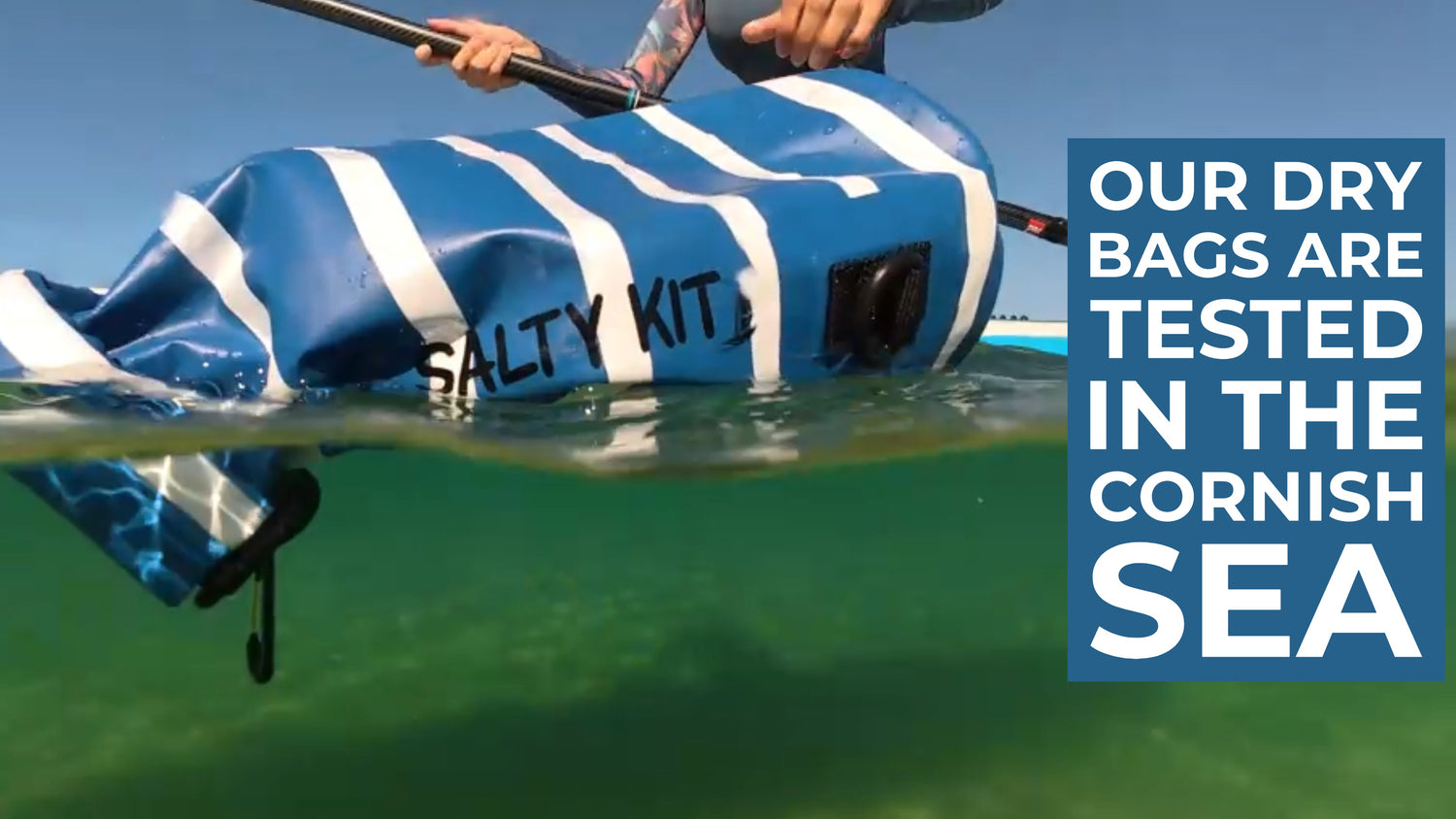 A blue and white striped dry bag labeled 'SALTY KIT' floating in clear greenish water, with a paddle visible and a blue text box on the right reading 'OUR DRY BAGS ARE TESTED IN THE CORNISH SEA'.