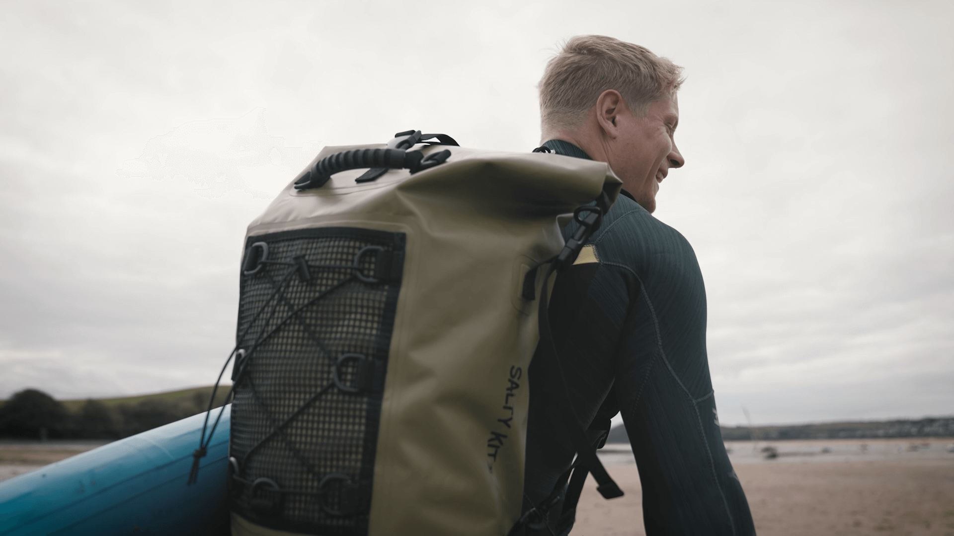 man walking on the beach with a paddleboard and wearing the 30 litre olive salty kit backpack